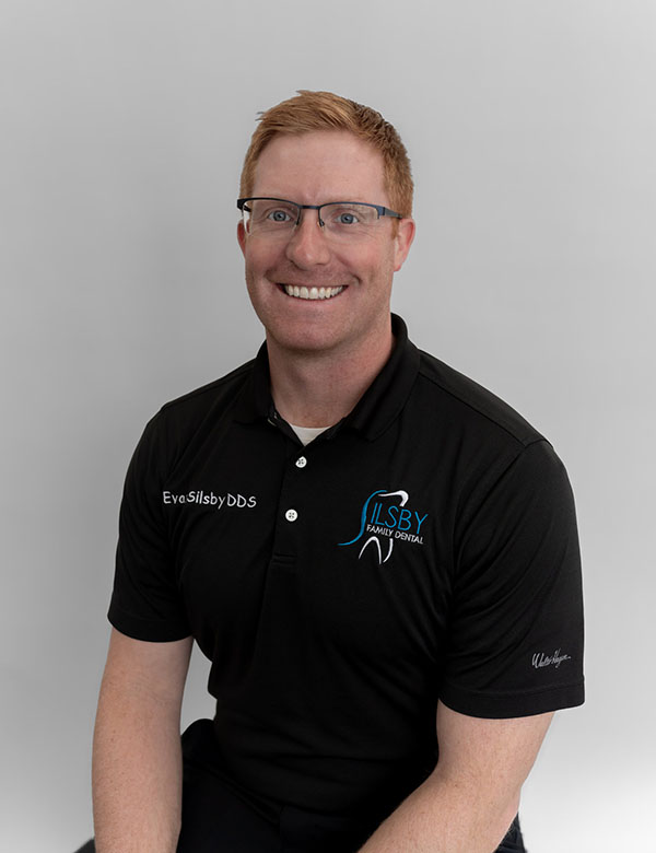 The image shows a smiling man wearing glasses, posing for a portrait with a white background. He is dressed in a black shirt with a logo on the left chest area and a dark-colored polo shirt underneath, along with a light-colored cap.