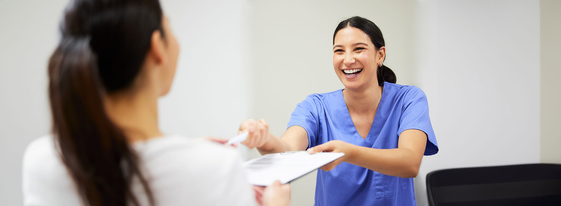 This image shows a woman in a white coat standing behind a desk with her hands on a clipboard, smiling and engaging with someone who appears to be handing her something there s another person in the foreground looking at them both.