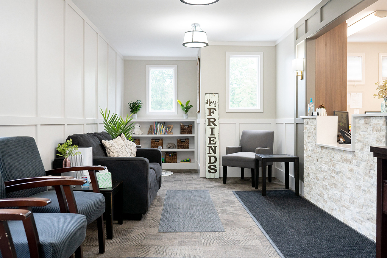 The image shows a modern interior of a waiting area with a clean and organized design, featuring seating, a reception desk, a sign, and a decorative wall.