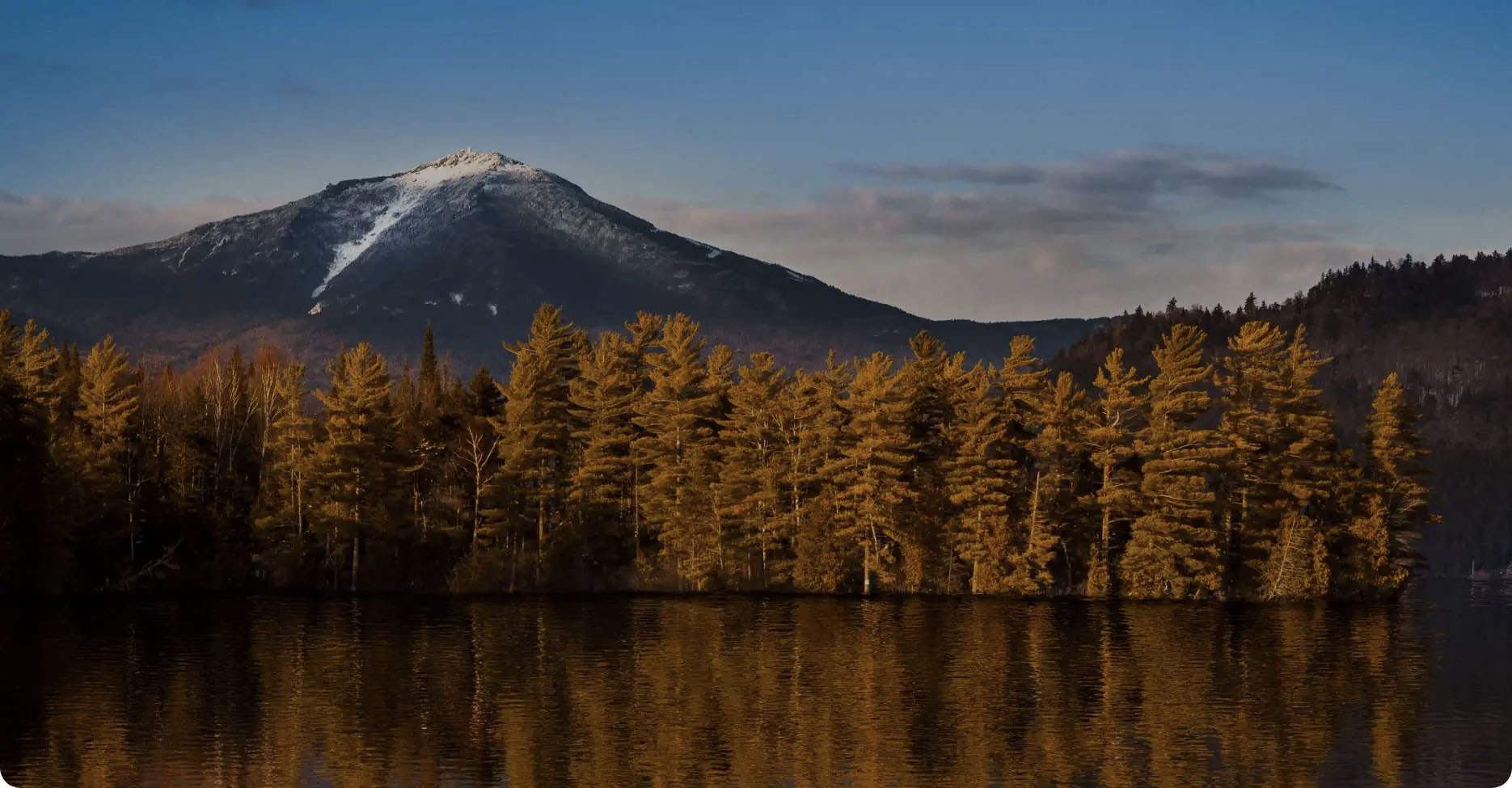 This image features a serene landscape with a mountain, trees, water, and a clear sky, showcasing a natural setting at sunset.
