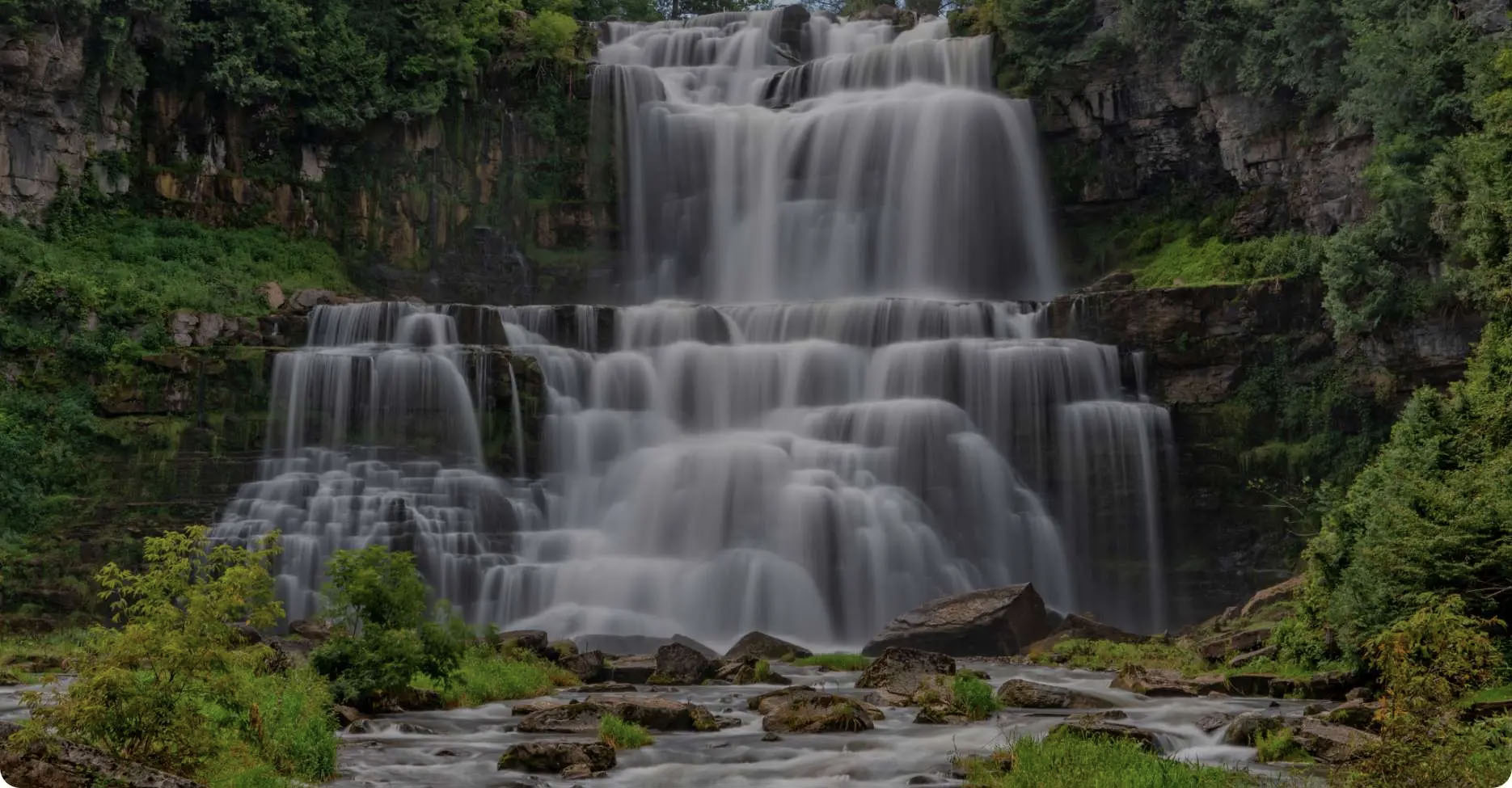 The image shows a majestic waterfall cascading down a cliff face, surrounded by lush greenery and trees, with a clear blue sky above.