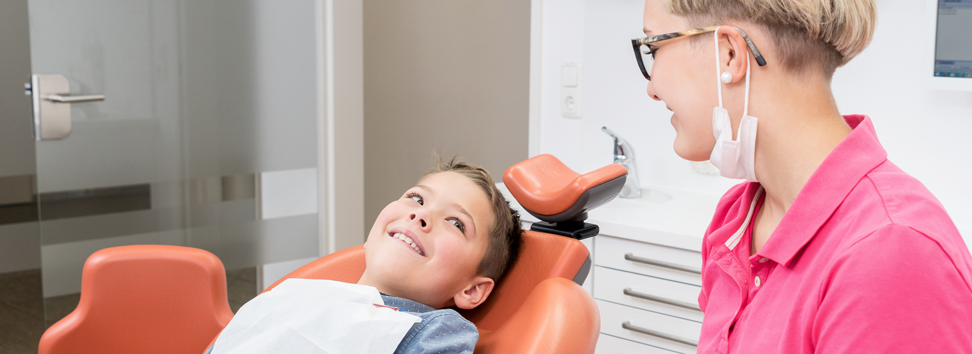 The image shows a dental office setting with a person sitting in the dentist s chair during an appointment, a dental professional standing nearby, and another individual taking a photo of the scene.