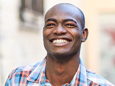 The image shows a smiling man with short hair, wearing a blue plaid shirt, standing outdoors against a backdrop of a building.